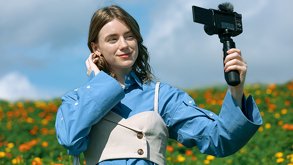 This is an image of a young woman wearing a blue-and-white jacket taking a selfie with the Canon PowerShot V1 outdoors, with orange and yellow flowers and a blue sky in the background.