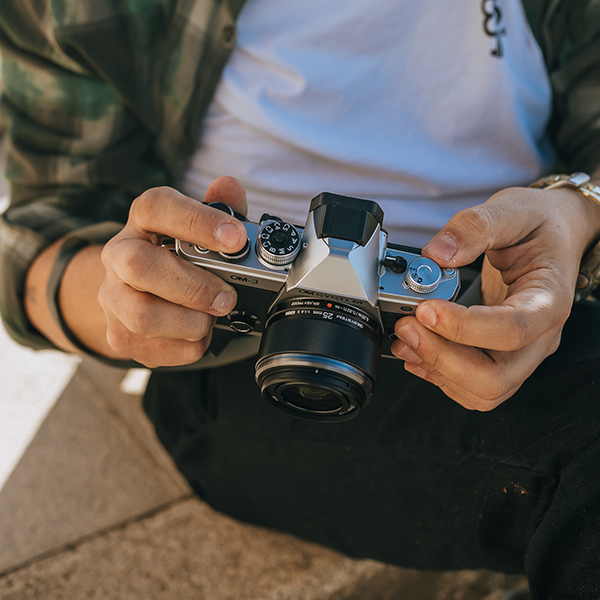 This is a photo of a photographer's hands holding OM SYSTEM OM-3 camera with the M.ZUIKO DIGITAL 25mm F1.8 II lens attached to the camera body.