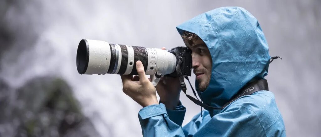 A photographer in a blue hooded rain jacket holding a camera with an OM SYSTEM M.Zuiko Digital ED 50-200mm f/2.8 IS PRO.