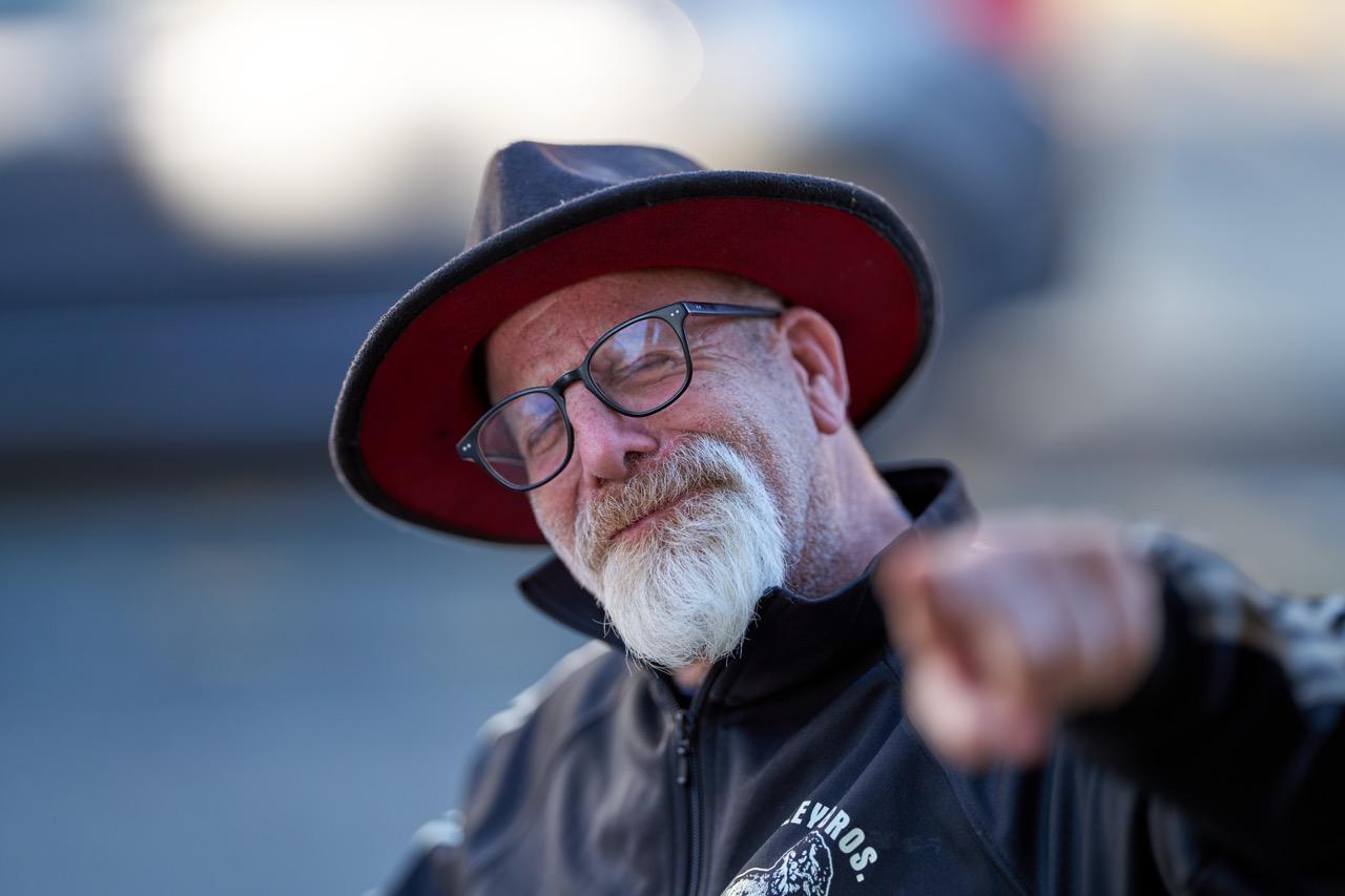 A man with a white beard, large hat, and glasses, points at the camera. 