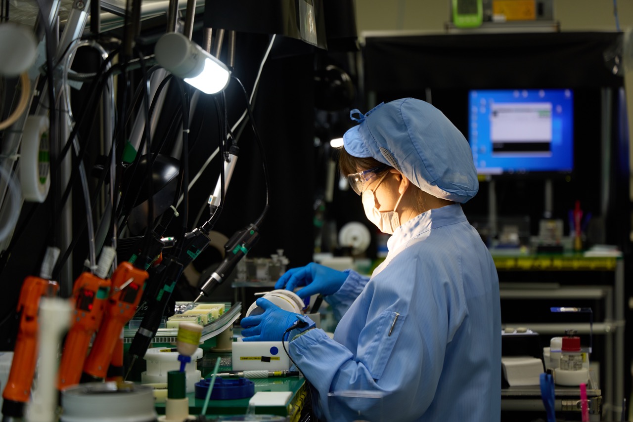 A woman stands at a factory assembly table constructing a Canon lens. 