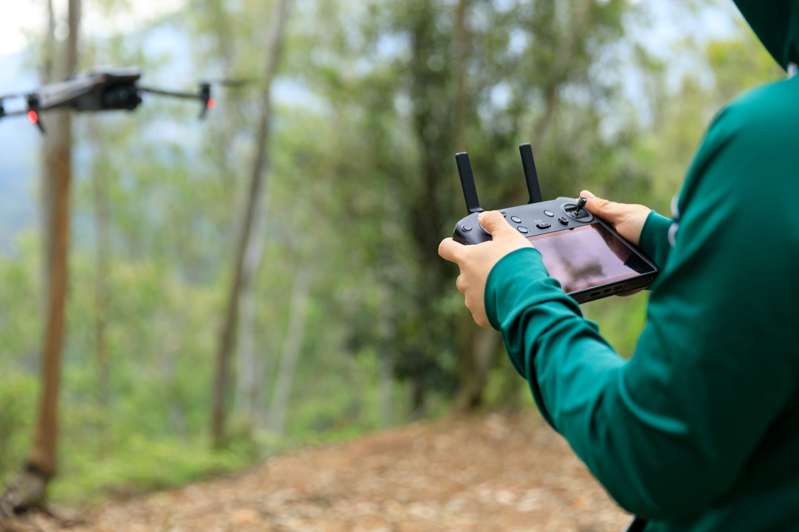 Side view of a person in a forrect holding the controller for a drone, while the drone hovers nearby. 